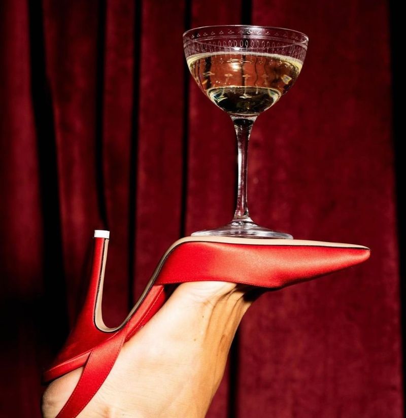 Un pied en escarpin rouge portant une coupe de champagne en équilibre, devant un rideau de velours rouge